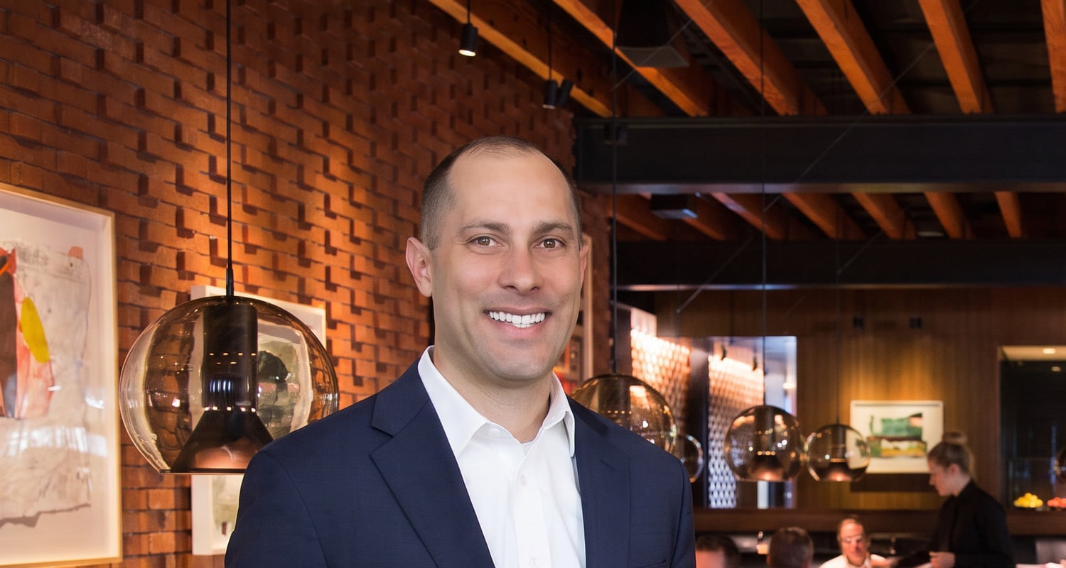 Jon Peck in a navy suit in a brick-walled restaurant dining room